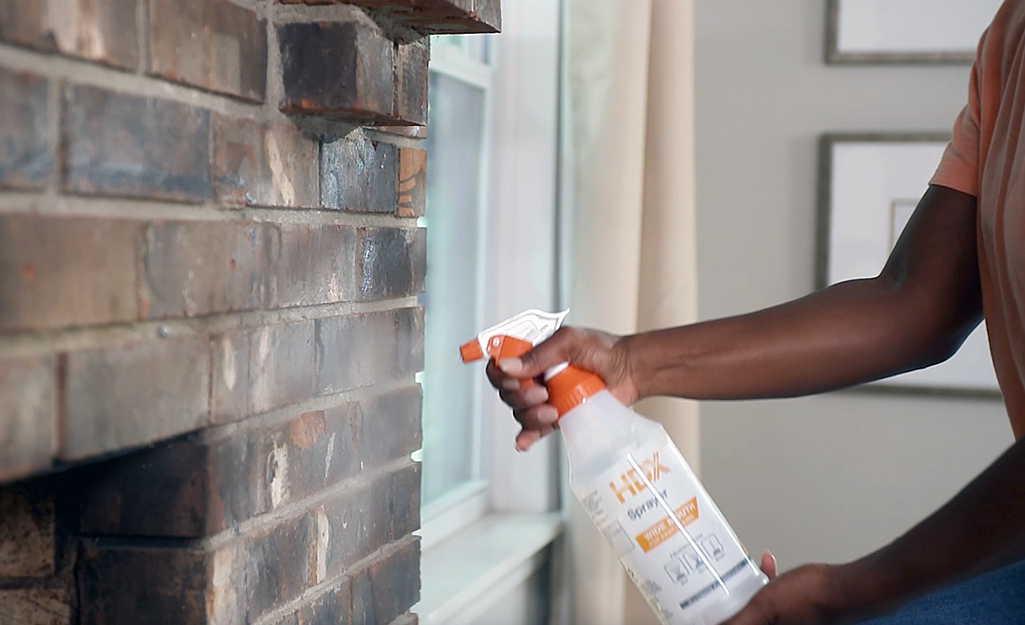 A person sprays a cleaning solution to a brick fireplace wall.