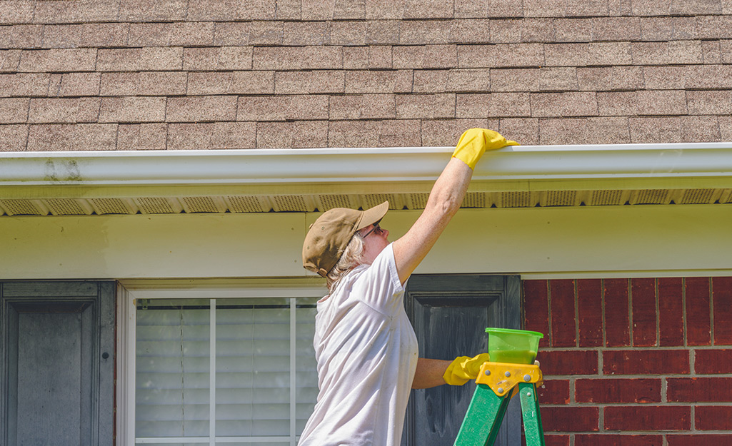 A person on a ladder cleaning debris from a gutter..