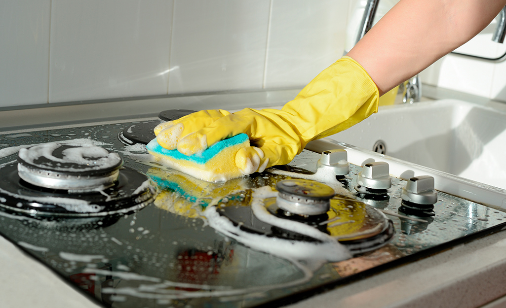 A person wearing kitchen gloves cleans a stove top with a sponge.