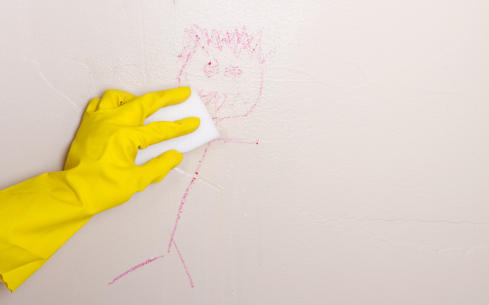 A person wearing gloves uses an erasing pad to clean a crayon drawing from a wall.