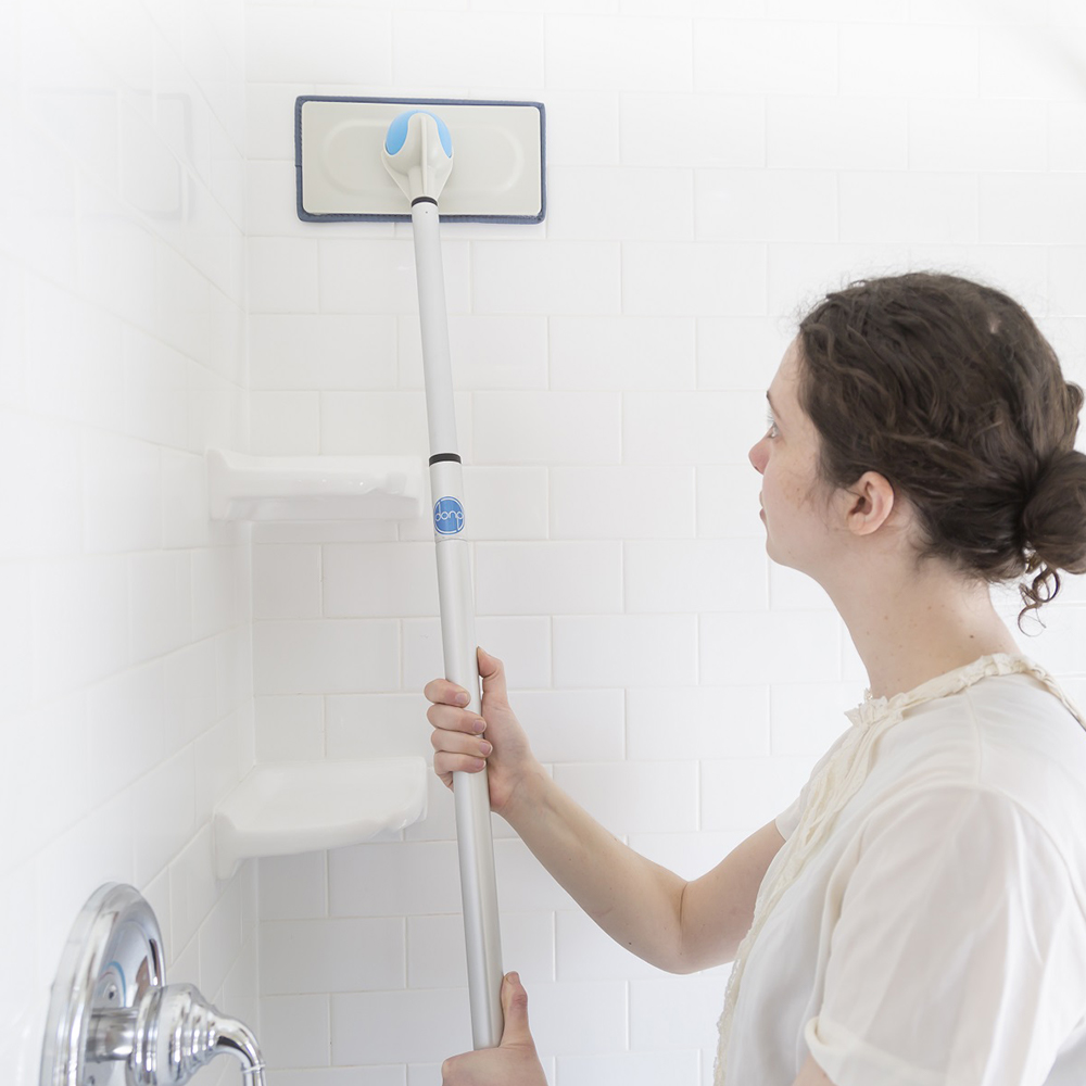 A woman uses a long-handled cleaning tool to wipe down a shower wall.