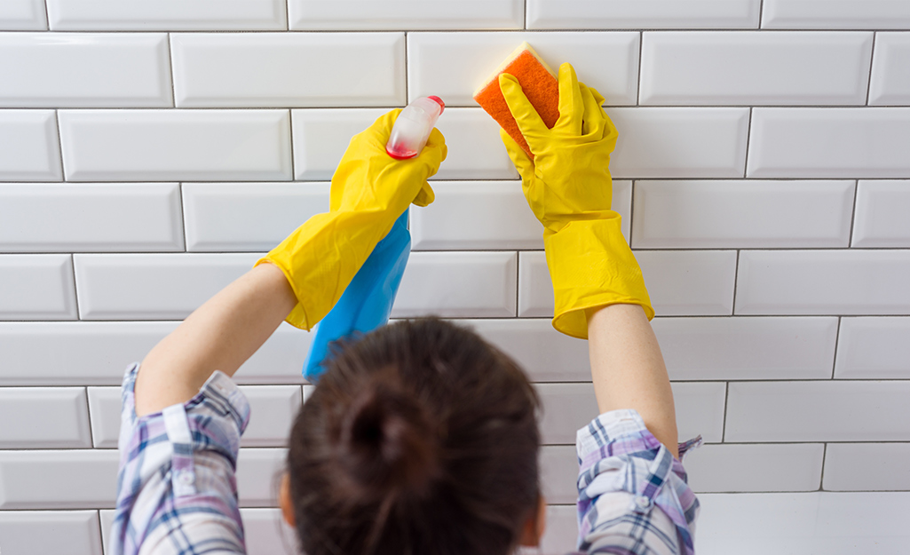 A person sprays a cleaning solution onto tile and wipes it with a sponge.