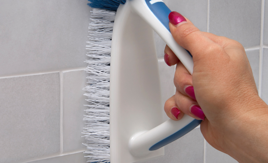 A person uses a scrub brush to clean shower tiles and shower grout.