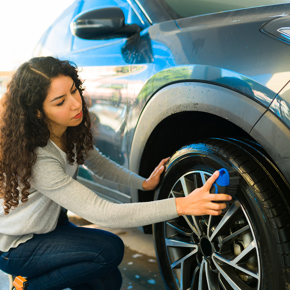 A woman uses a brush to clean the tire of an SUV.