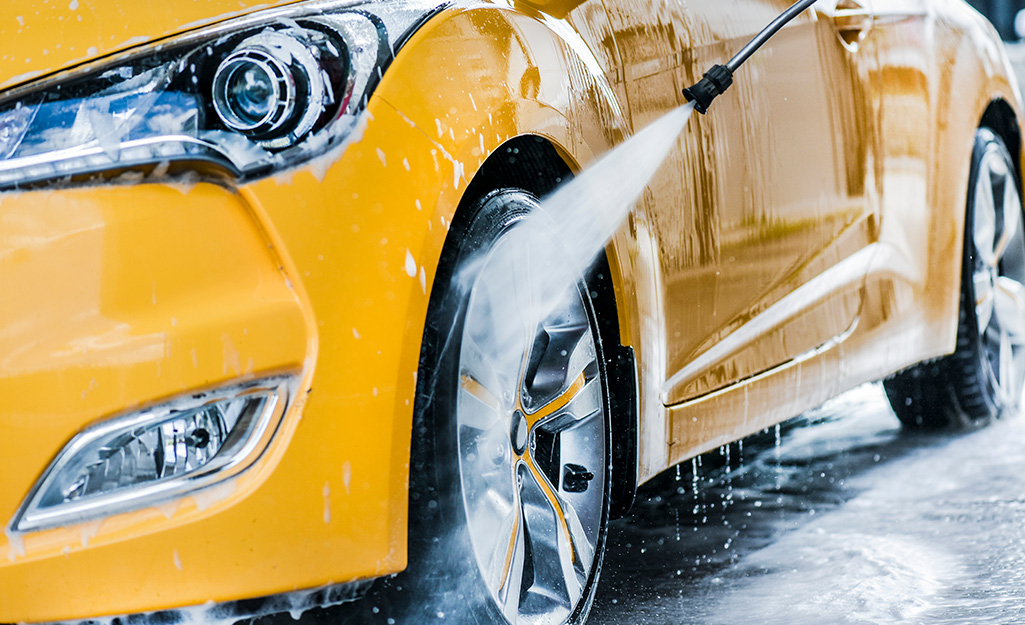 A person washes a tire on a yellow car with the spray nozzle of a hose.