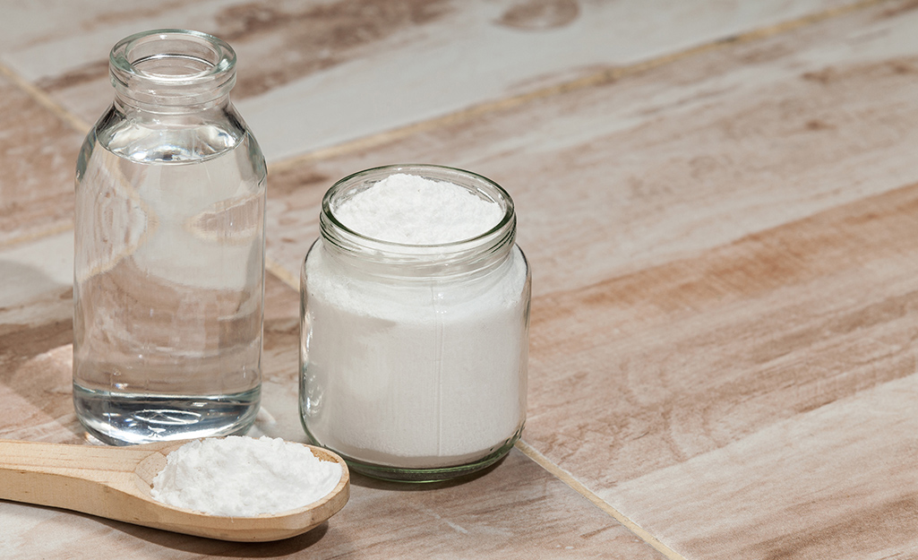 Glass jars of baking soda and vinegar sit with a wooden measuring spoon on a vinyl floor.