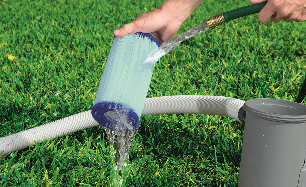 A person holds a pool filter and rinses it off using a hose. Water runs off the filter and onto grass.