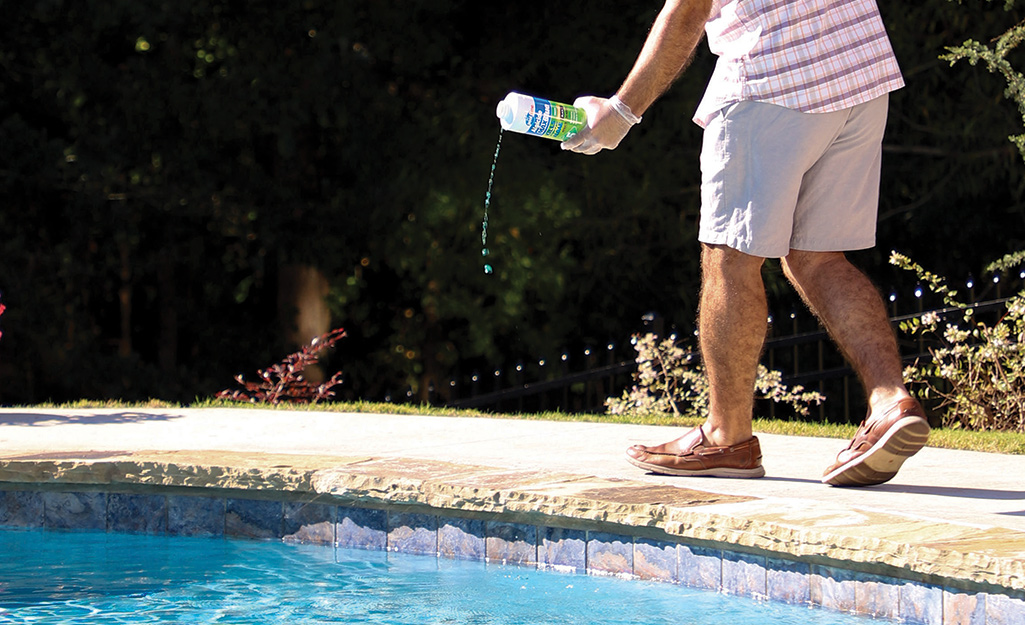 A person sprinkles chemicals into pool water as they walk beside a pool.