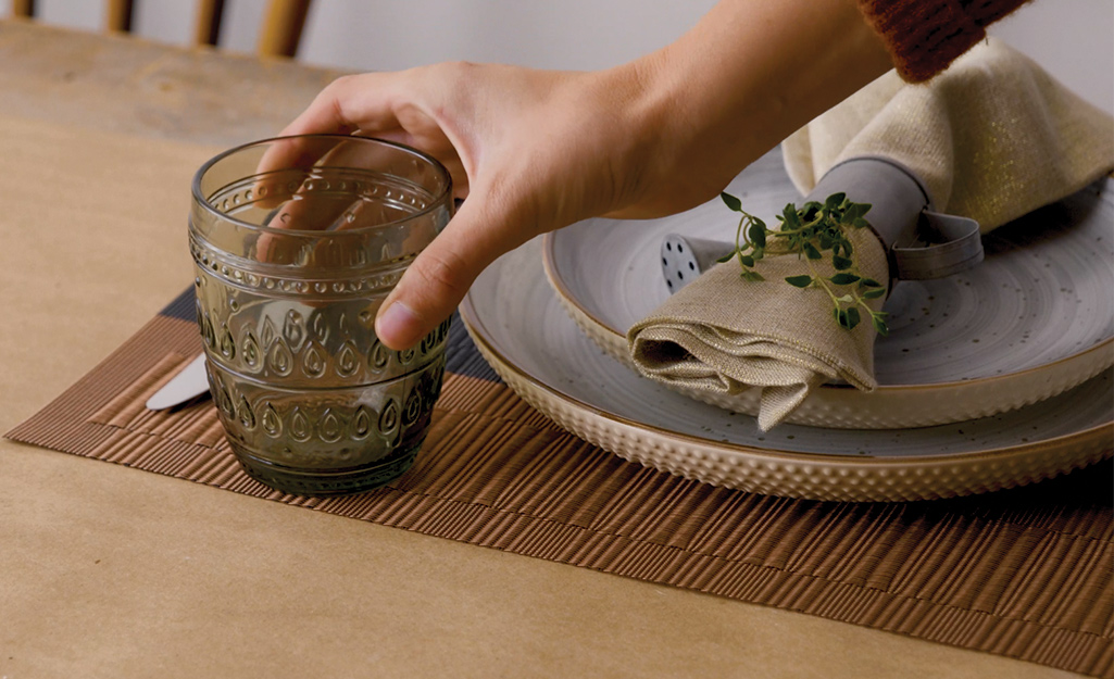 A person setting a table with a glass, plates and napkin rolls.