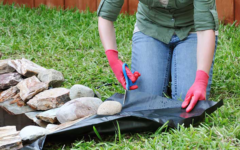 A woman places water plants into a small backyard pond surrounded by paver stones.