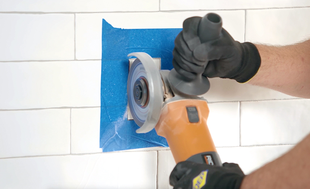 A person uses an angle grinder to cut a piece of tile from a white bathroom wall.