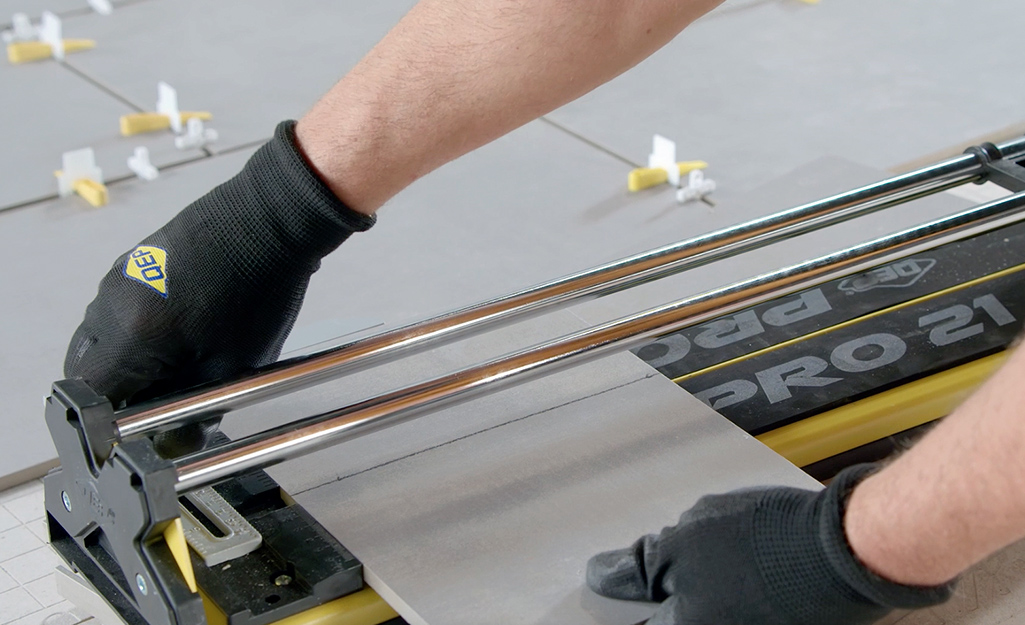 A person using a manual tile cutter to make a straight cut on a piece of tile.