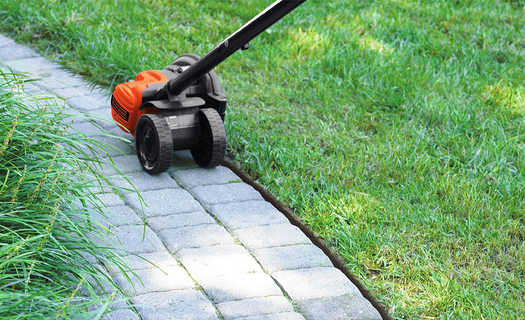 A person using an electric edger to trim the grass beside a paver walkway.