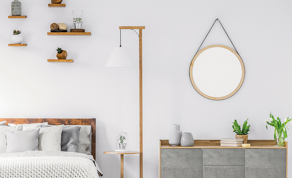 A circular mirror hangs from the wall above a gray dresser that’s next a floor lamp, night table and bed.