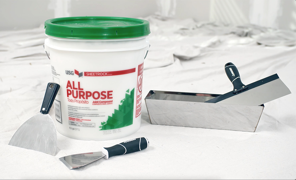 A bucket of drywall compound, drywall mud knives and a mud pan sit on a white drop cloth.