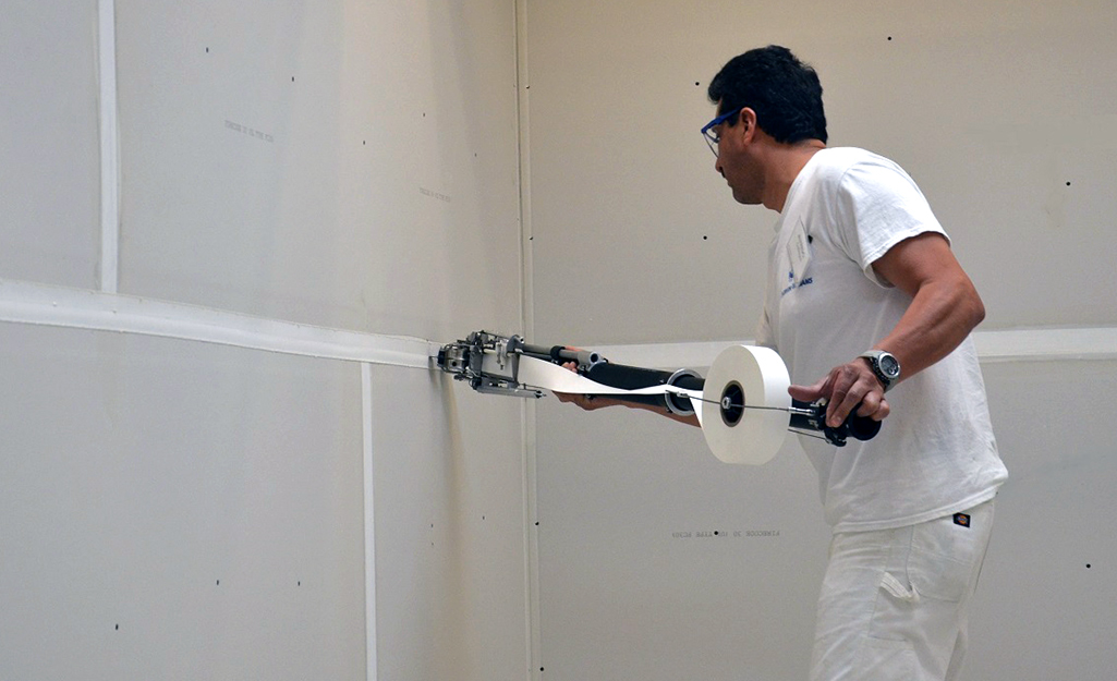 A man unrolls paper drywall tape onto the seam between two sheets of drywall.