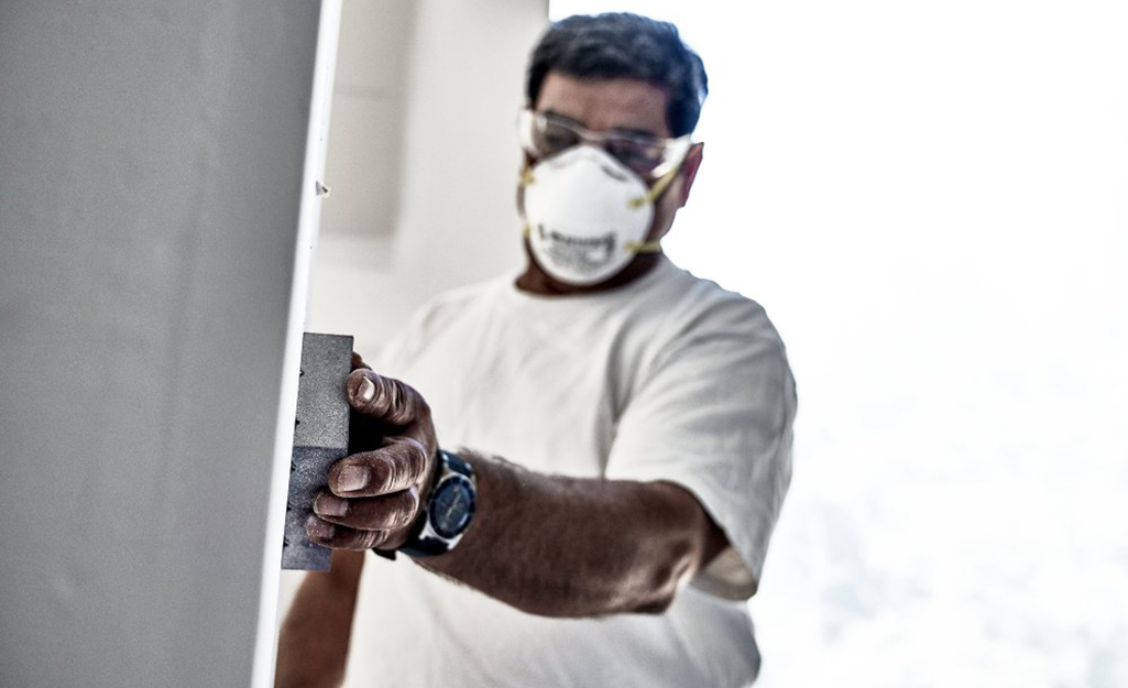 A man wearing a safety mask sands drywall with a sanding block. 