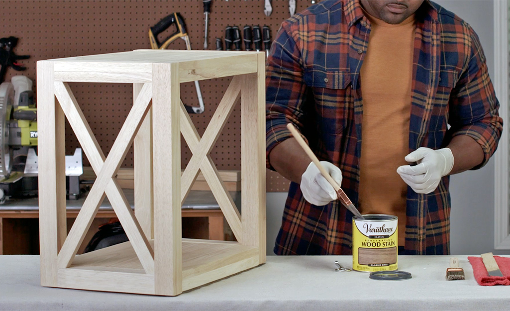 A person wearing gloves dips a brush into a can of wood stain next to an unfinished wood project.