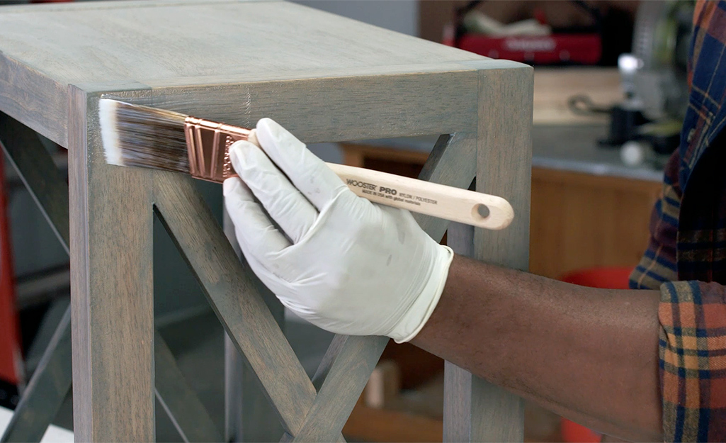 A person wearing a glove applies stain to wood with a brush.