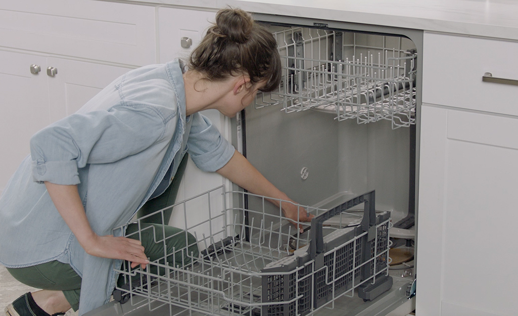 A woman wearing a blue shirt kneels down and reaches into the bottom of an open dishwasher with the bottom rack pulled out. 