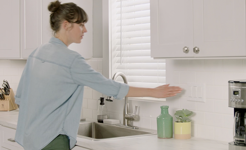A woman wearing a blue shirt stands at a kitchen sink and extends her arm to turn on a switch to the garbage disposal.