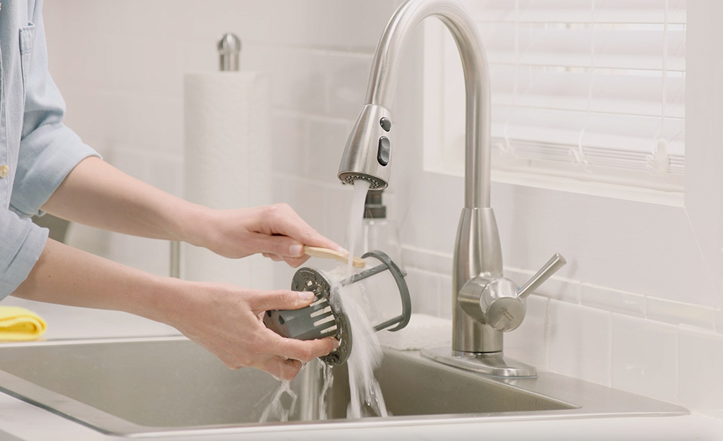 A person uses a toothbrush to clean a dishwasher filter under the running water of a stainless steel kitchen sink.