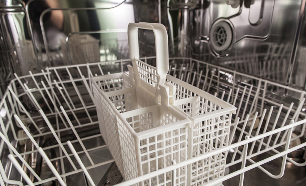 A white basket for silverware stands in the middle of the bottom rack inside a dishwasher with shiny metal walls.