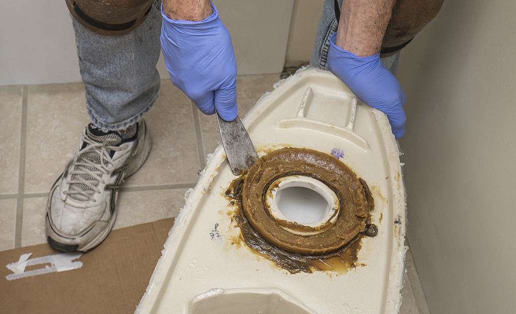 A person scraping off the wax ring on the bottom of a toilet.