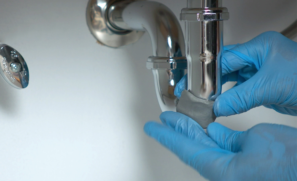 A person applies epoxy putty to the pipe beneath a sink. 