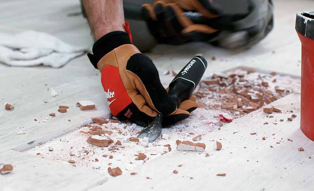 A person wearing gloves uses a hammer and a chisel to remove a piece of damaged tile from a floor.