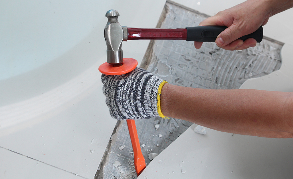 A person wearing gloves uses a hammer and a chisel to remove a piece of damaged tile from a floor. A person wearing gloves uses a hammer and a chisel to remove a piece of damaged tile from a floor.