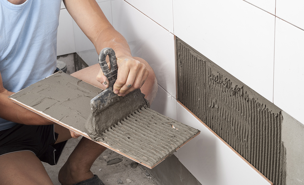 A person applies thin-set mortar to the back of a new tile before placing it on a tiled wall. A person applies thin-set mortar to the back of a new tile before placing it on a tiled wall.