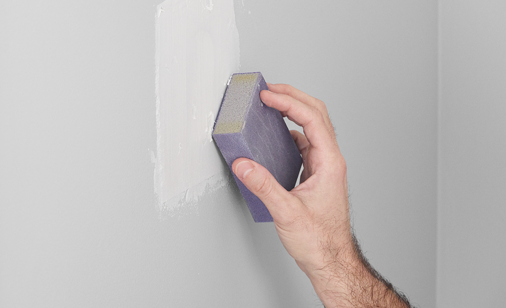 A person uses a sanding sponge to smooth a spackled patch on a wall.