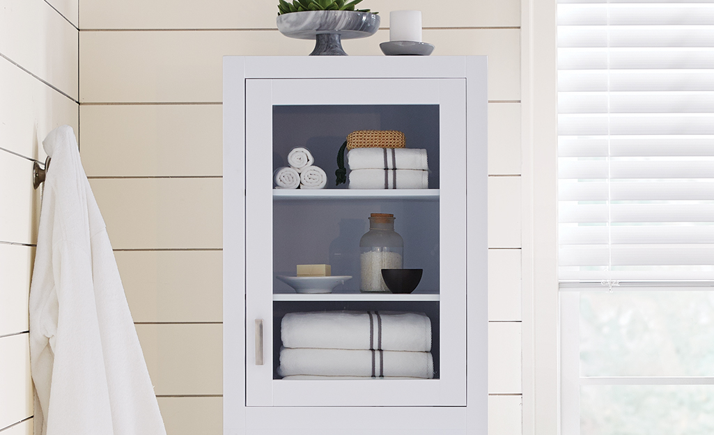 White towels is various sizes are stacked in a white cabinet with a glass door.