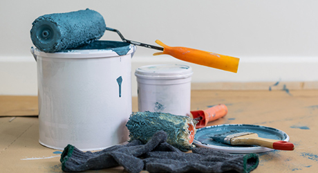 A paint roller soaked in blue paint sits on top of white paint bucket next to paint brush and rags.