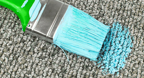 A paint brush soaked in blue paint lays next to dried paint stain on carpet.