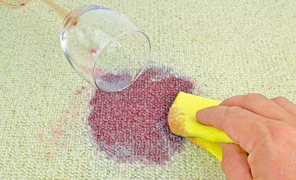 A person blots red wine spilled from a glass onto a carpet.