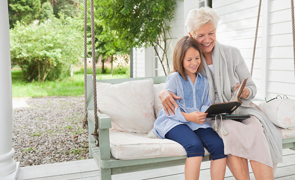 A woman with gray hair shares a book with a girl while they sit in a porch swing. 