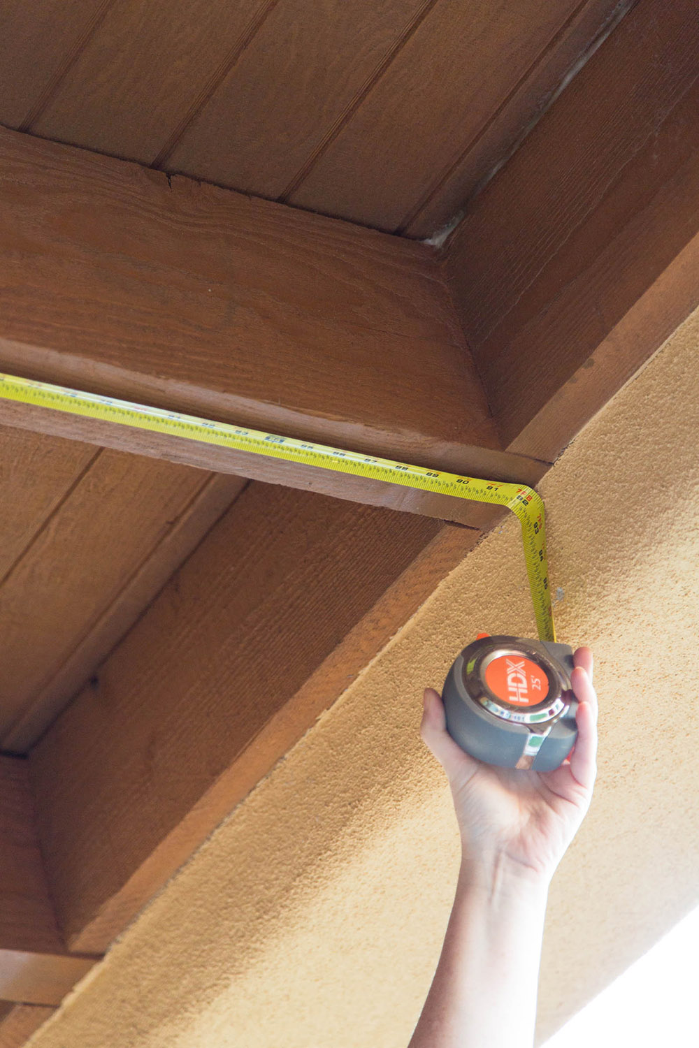 A tape measure is held up against a wooden ceiling beam and tan wall.