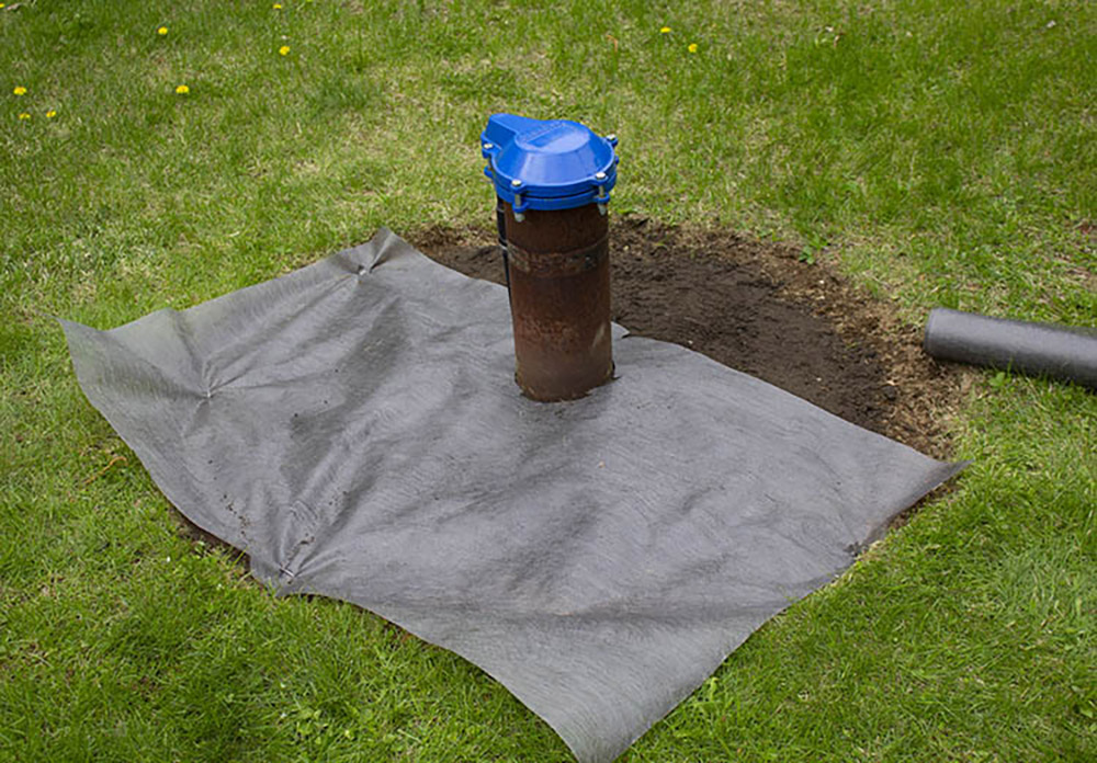 A square piece of landscape fabric covering one side of the ground next to an exposed wellhead.