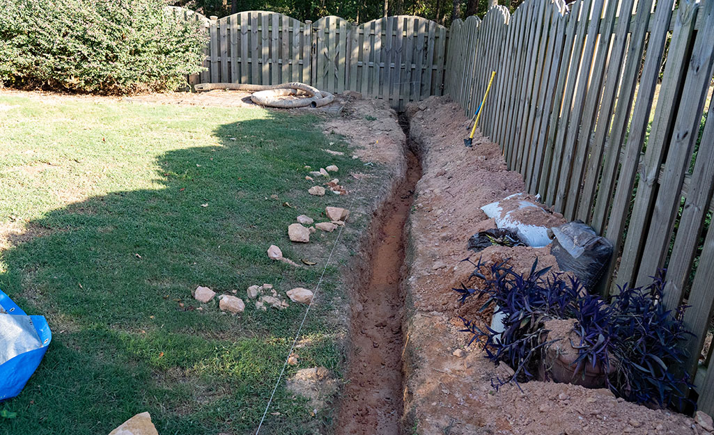 A French drain trench dug along the edge of a fenced-in yard.