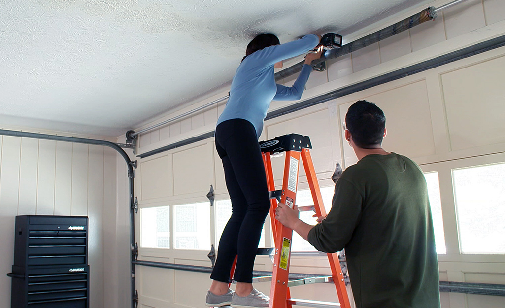 A man holds a ladder as a woman stands on it and reaches above a garage door to attach a bracket to the wall.