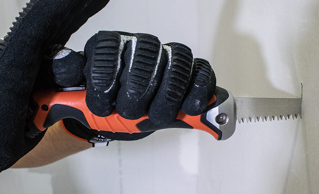 Someone wearing a work glove and a drywall saw to cut into a wall. Someone wearing a work glove and a drywall saw to cut into a wall.