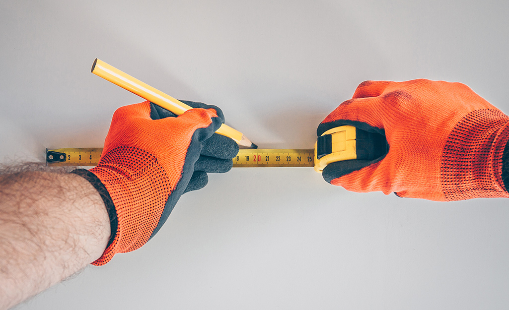Someone wearing orange work gloves using a carpenter's pencil and tape measure to mark a wall. Someone wearing orange work gloves using a carpenter's pencil and tape measure to mark a wall.