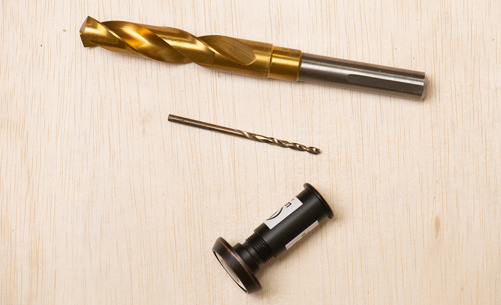 Two drill bits and a door viewer, or peephole unit, on a piece of plywood.