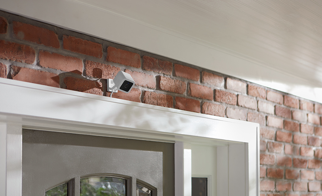 A security camera is installed on a brick exterior wall above the front door to a house. A security camera is installed on a brick exterior wall above the front door to a house.