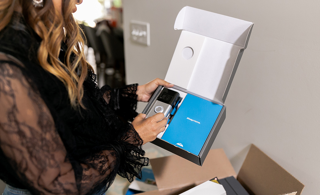 A woman wearing a shirt with a lacy black sleeve opens a box that contains a new security camera. A woman wearing a shirt with a lacy black sleeve opens a box that contains a new security camera.