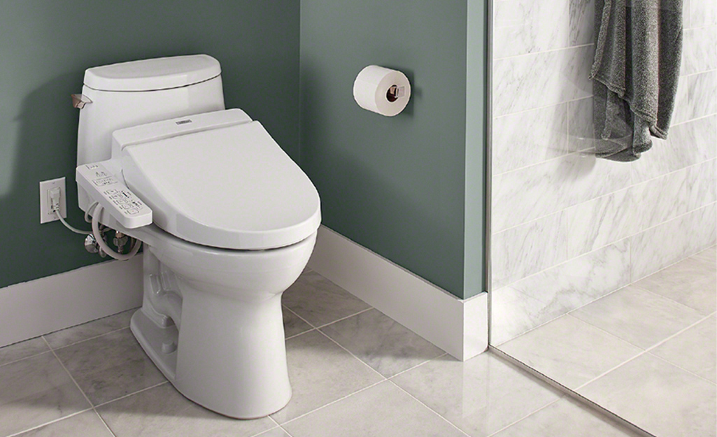 An electric toilet bidet seat connected to a bathroom wall outlet.