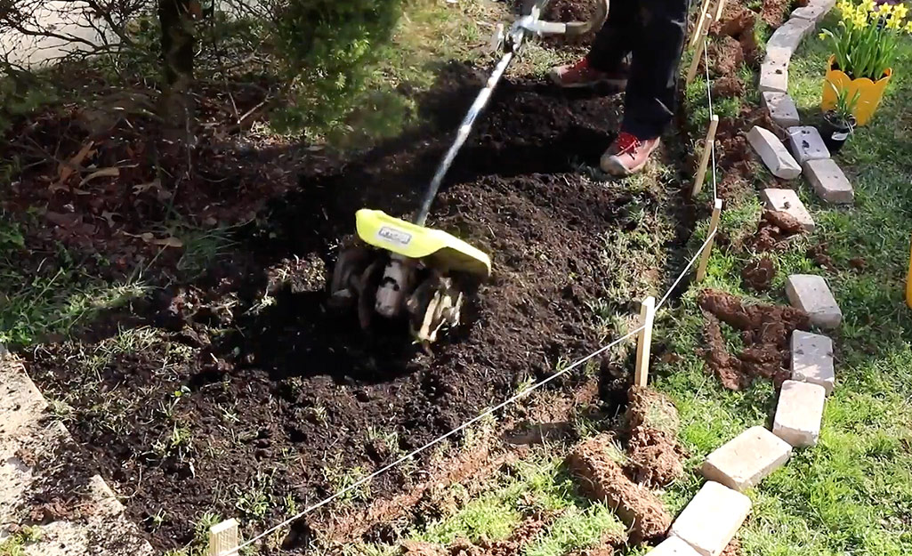 A person uses a cultivating tool to break up soil for a garden bed.