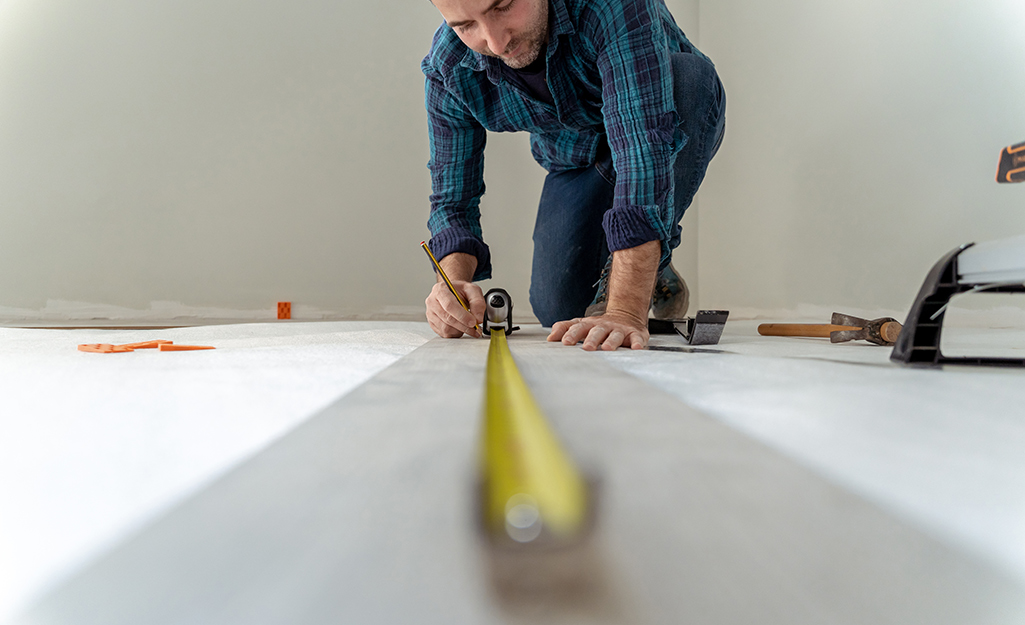 A tape measure extends in front of a man who kneels to measure the dimensions of a room before installing carpet.